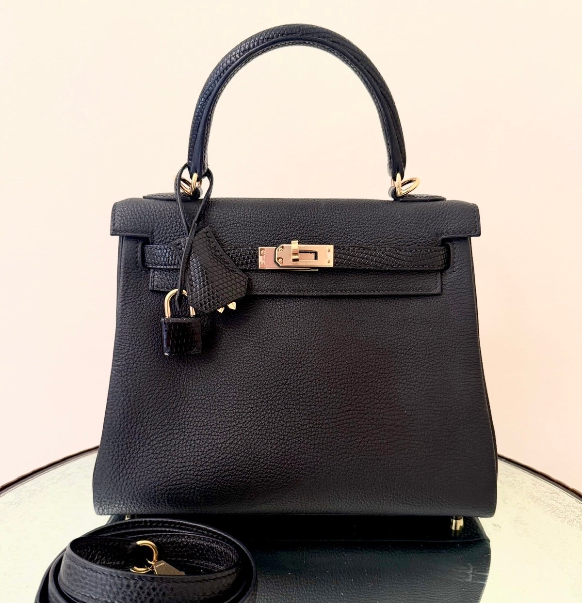 A black pebbled leather handbag with gold-tone hardware, featuring a top handle, front flap, clochette, and lock, displayed on a reflective surface with a matching strap in front.